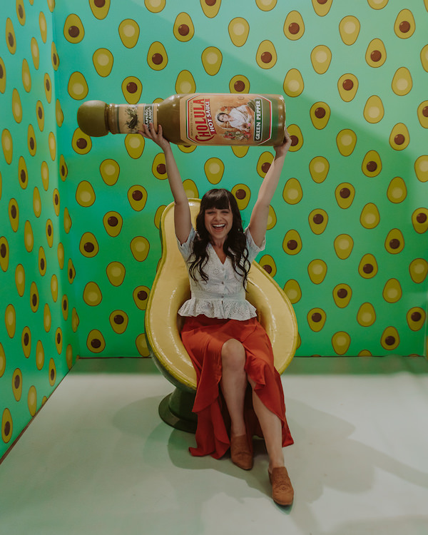 Woman sitting in avocado shaped chair holding oversized bottle of hot sauce