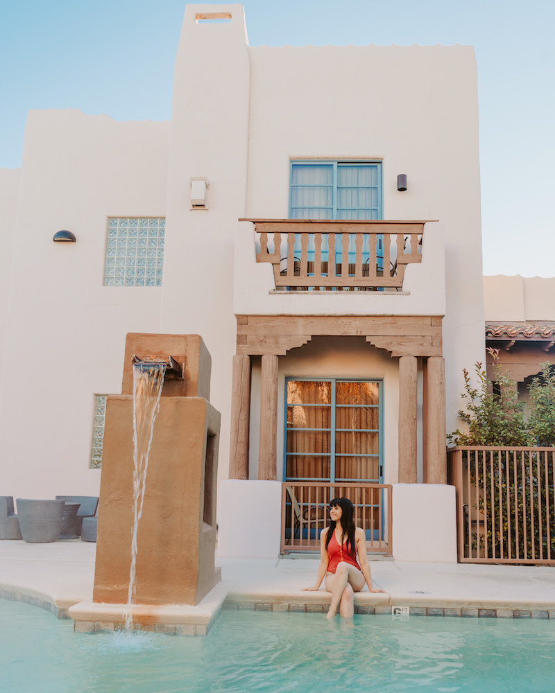 Woman in swimsuit sitting at edge of swimming pool in front of adobe building