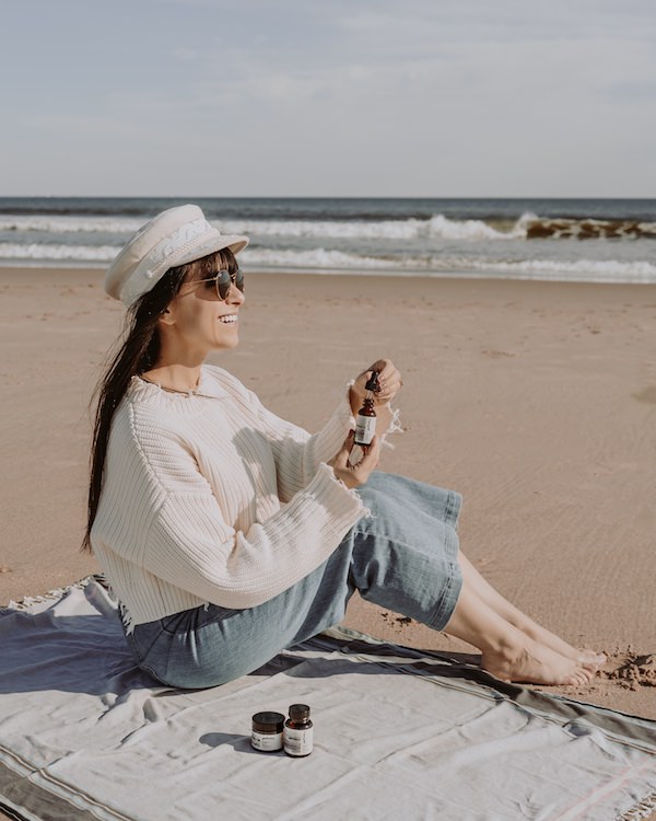 woman sitting on beach holding glass bottles of beauty products
