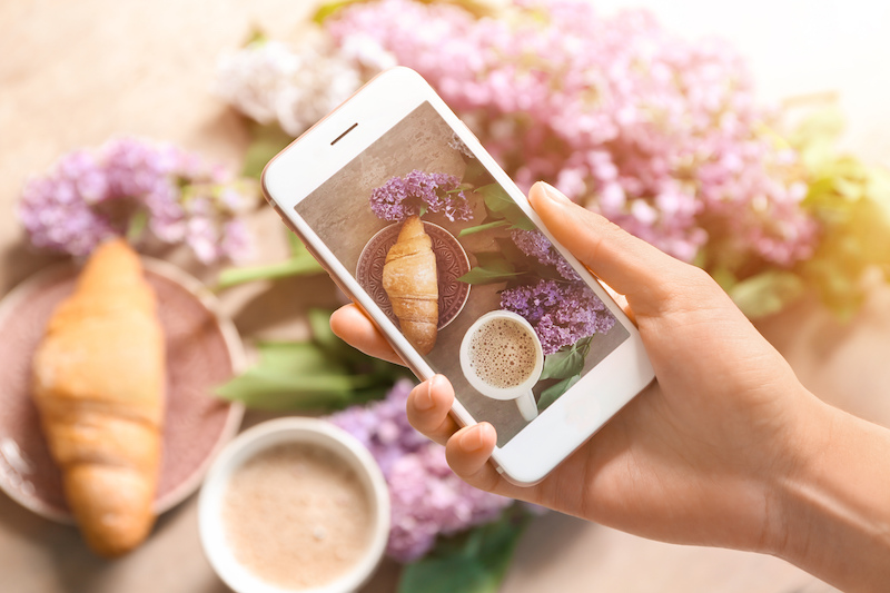 Woman's hand taking photograph of flowers, coffee and croissant with white iphone for the new Instagram algorithm of 2020
