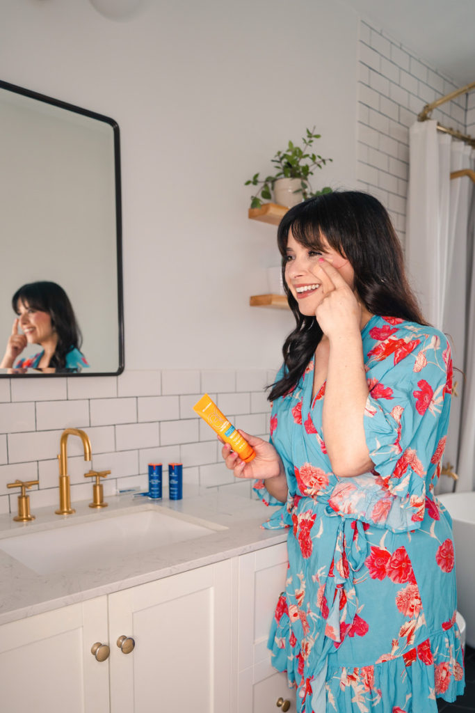 Woman in blue and red floral robe applying sunscreen from an orange tube to her face - MDSolarSciences Mineral Tinted Creme - in a white tiled bathroom