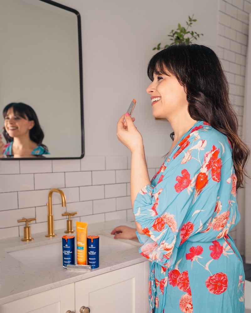 MDSolar Sciences review: woman wearing blue and floral robe applying lip balm in a white tiled bathroom as part of a winter skincare routine