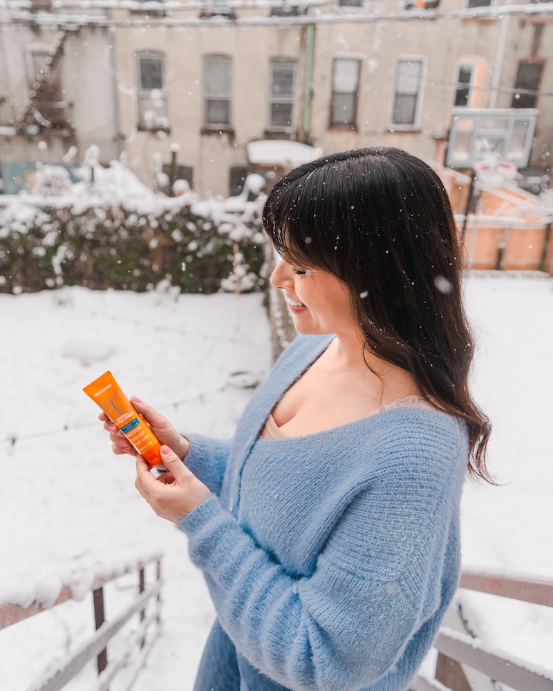 Woman wearing light blue sweater and holding an orange tube of MDSolarSciences Mineral Tinted Creme mineral sunscreen in a snowy backyard