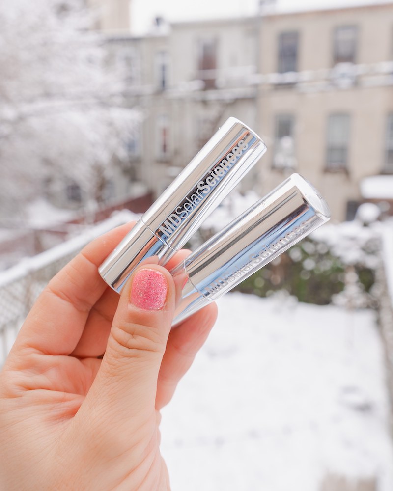 Woman's hand holding two silver tubes of MDSolarSciences Hydrating Sheer Lip Balm SPF 30 against an outdoor snowy backdrop
