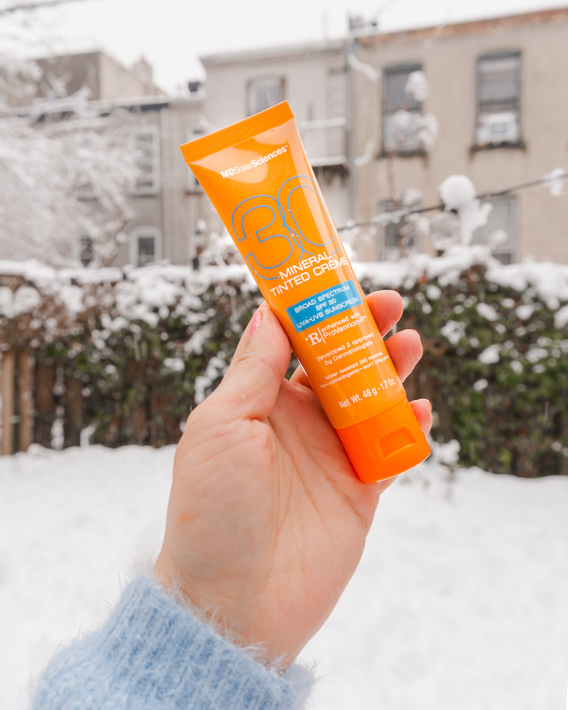 Woman's hand holding orange tube of MDSolarSciences Mineral Tinted Creme mineral sunscreen against an outdoor snowy backdrop