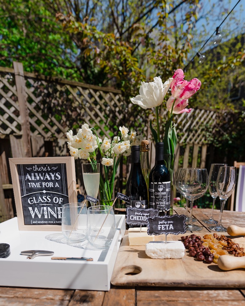 A close up of a wooden table. outside in an urban backyard. with wine bottles, wine glasses, and a cheese platter 
