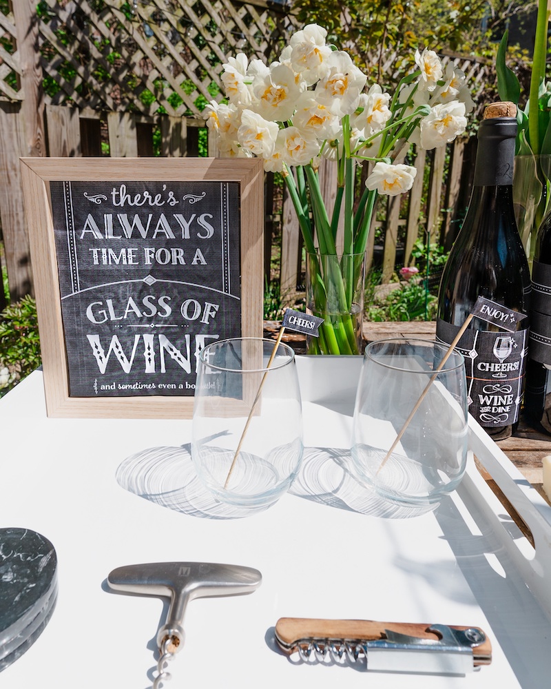 A close up photo of party decorations by Hostess With the Mostess Printables. A white tray with wine glasses and corkscrews and a black and white sign announcing a wine tasting event.