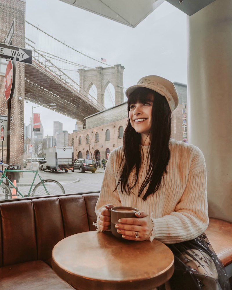 Woman wearing beige newpaper boy hat an beige sweater smiling and holding a coffee. She is sitting in a Brooklyn coffeeshop where the window shows a view of the Brooklyn Bridge. 