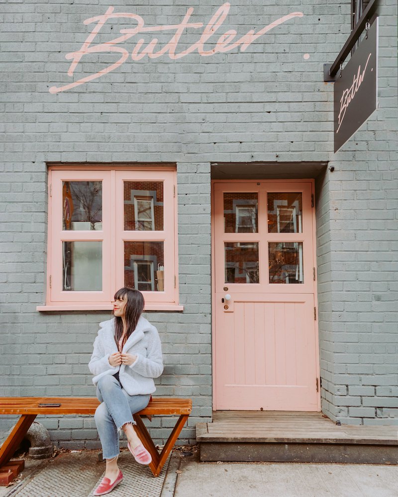 The grey and pink exterior of a coffee shop in Brooklyn. A brunette woman in a fake fur coat and jeans is sitting on the bench in front.