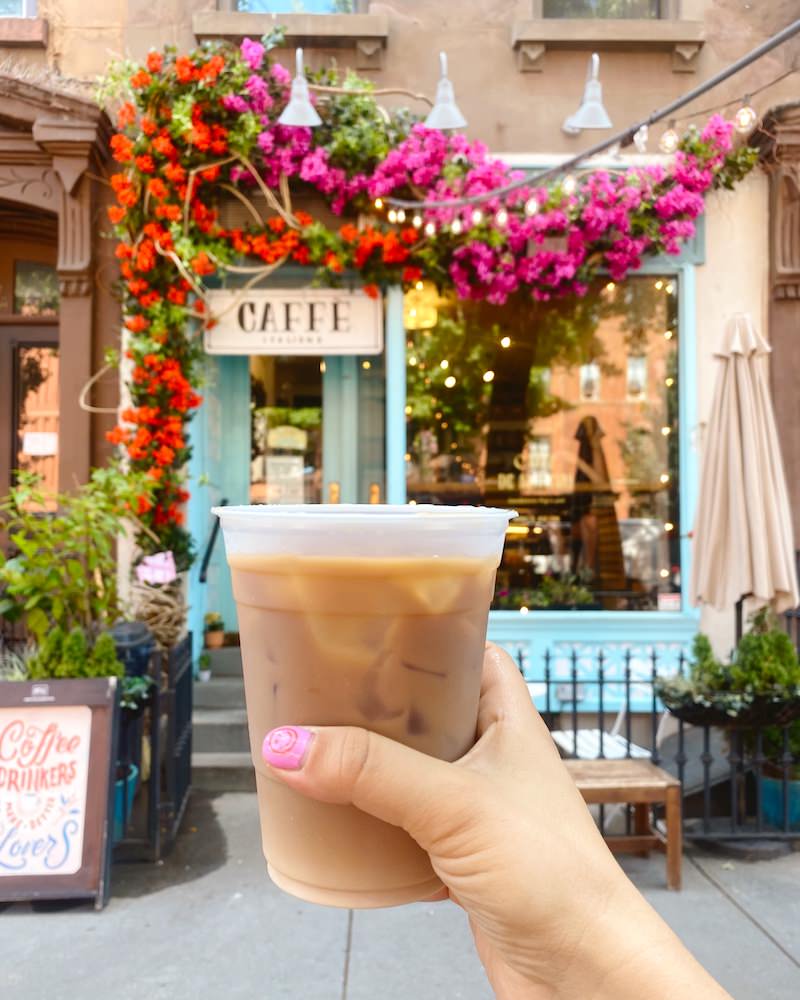 A woman's hand holding an iced coffee with a Brooklyn coffee shop's facade in the background. The coffee shop is painted turquoise and has pink and red flowers draped across the exterior.