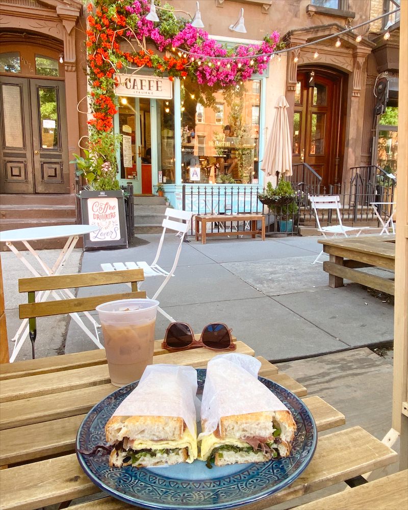An egg sandwich on a blue plate on a wooden table. The backdrop is the exterior of a coffee shop in Brooklyn.