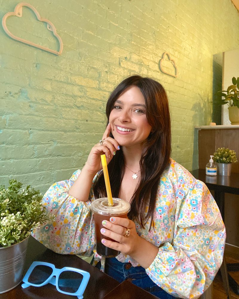 Brunette woman in multicolored floral top smiling at the camera and holding a boba / bubble tea while sitting in a Brooklyn coffee shop with seafoam green walls.