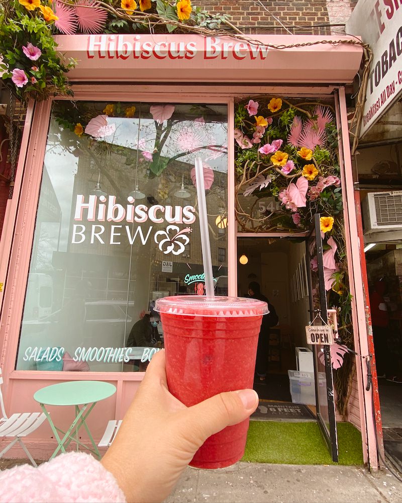 A woman's hand holding a bright pink smoothie in front of a pink coffee shop facade in Brooklyn.