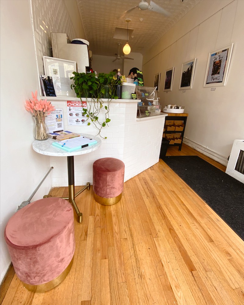 The interior of a small coffee shop in Brooklyn with wooden floors, a white counter, a small white table and pink velvet stools.