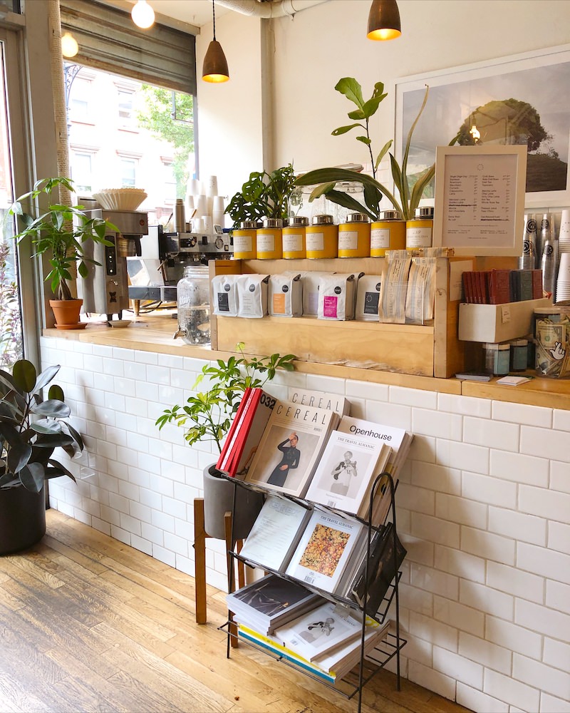 White tile and wooden counter of a Brooklyn coffee shop. The counter has coffee making products and green plants on it.