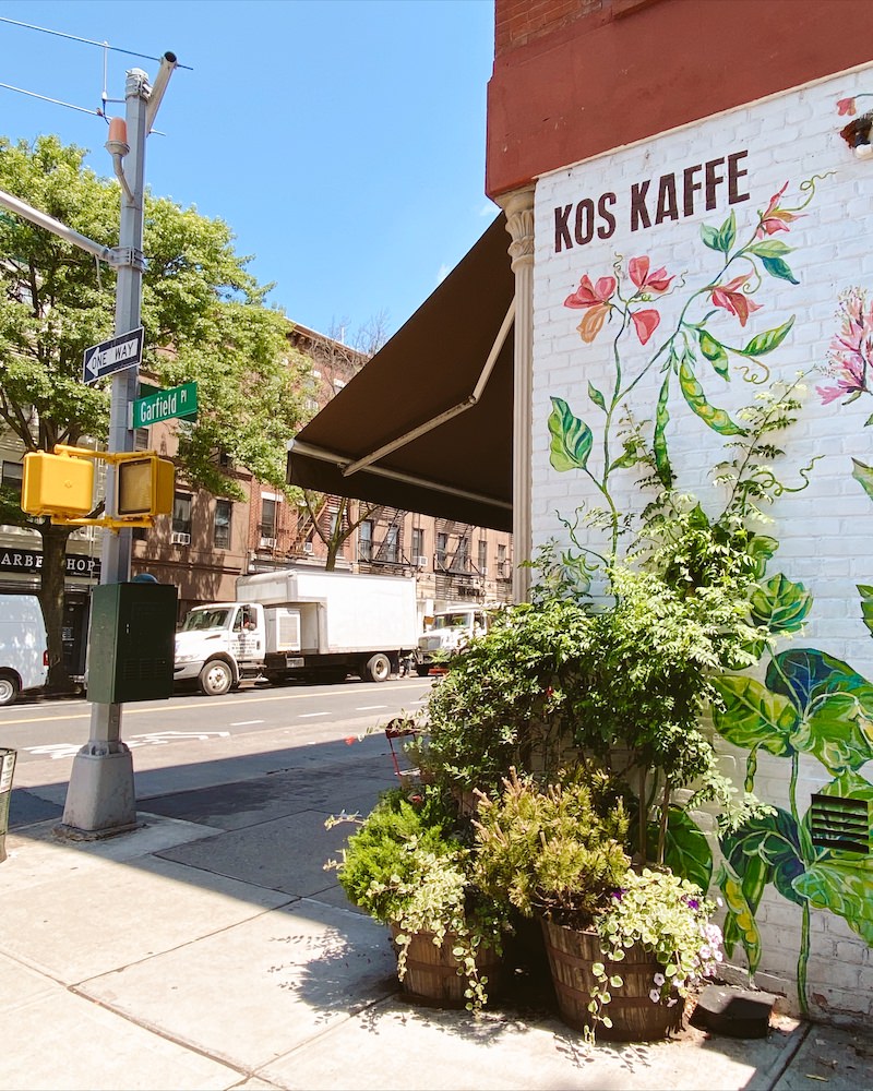 Exterior of a coffeeshop in Brooklyn with a black awning, and pink and green floral mural on a white brick wall.