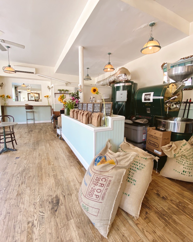 Interior of a coffee shop with roasting equipment, brown sacks of coffee beans, a hardwood floor and light blue counter.