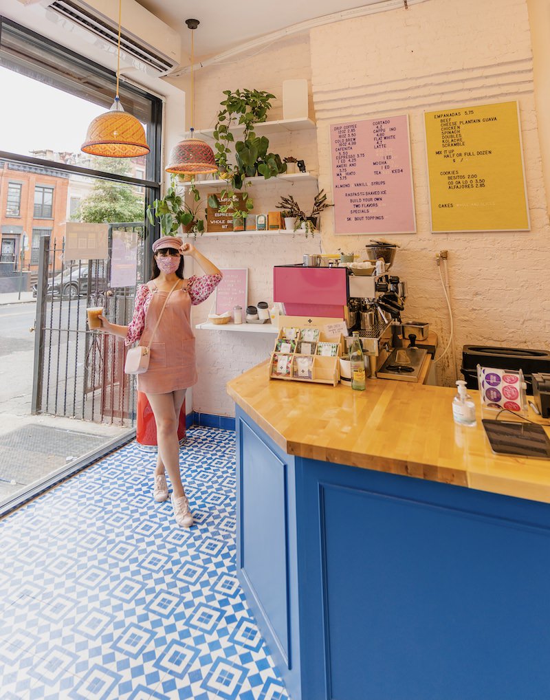 The interior of a colorful Brooklyn coffee shop that has a bright blue and wooden counter, bright pink espresso machine, pink and yellow letterboard menus and white shelves with green plants. A woman wearing pink is standing inside and holding a coffee.