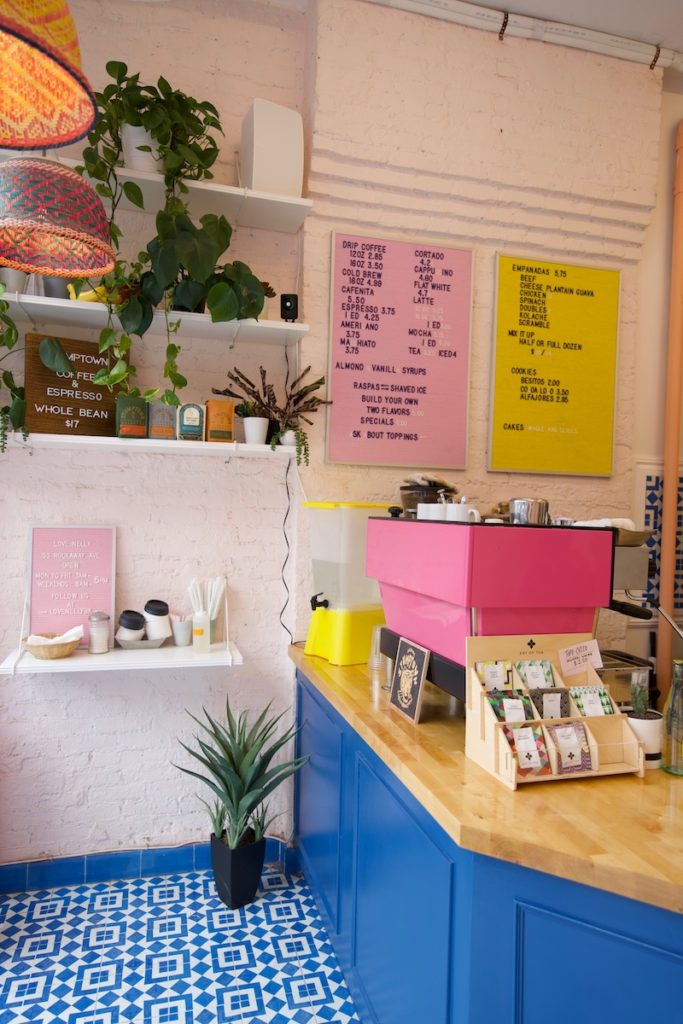 The interior of a colorful Brooklyn coffee shop that has a bright blue and wooden counter, bright pink espresso machine, pink and yellow letterboard menus and white shelves with green plants.