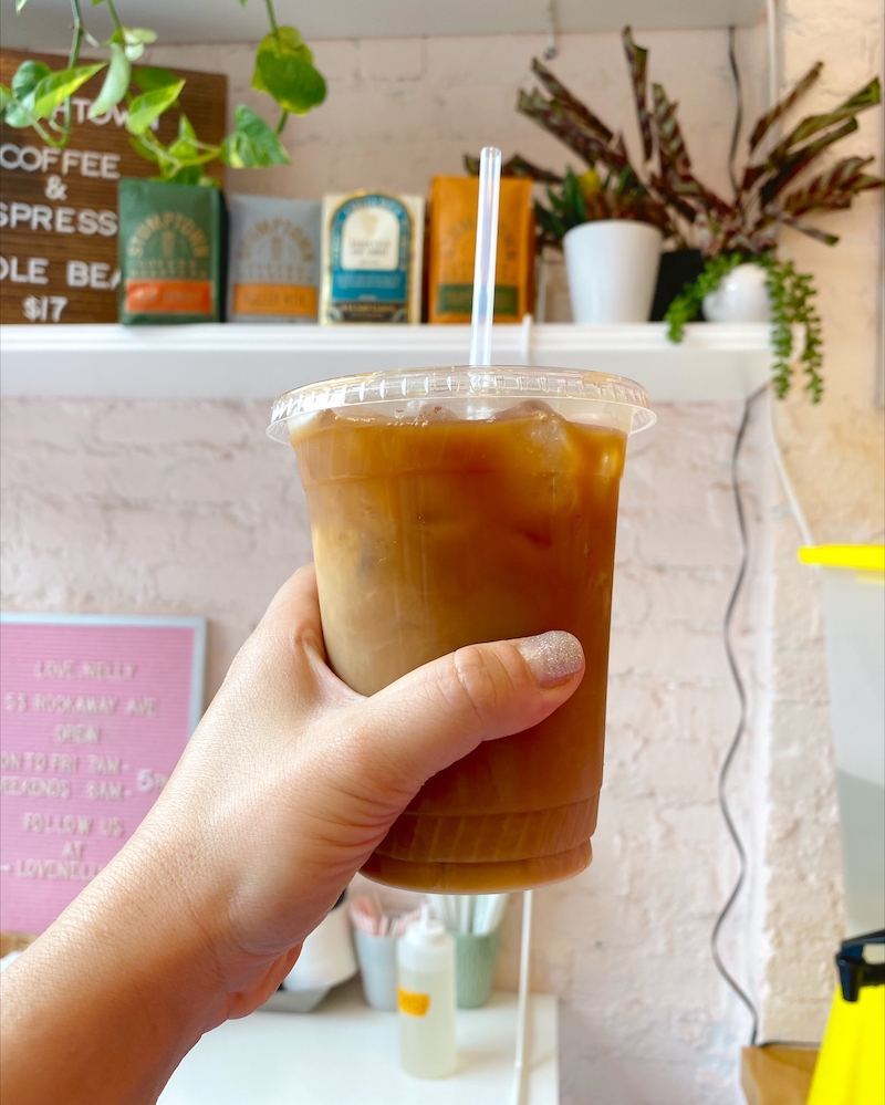 A woman's hand holding an iced coffee in front of a white shelf full of green plants.