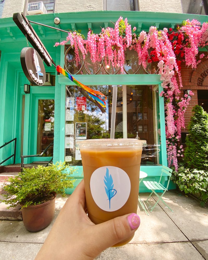 A close up photo of a woman's hand holding an iced coffee in front of a green facade of a Brooklyn coffee shop that is decorated with a pink floral arch.