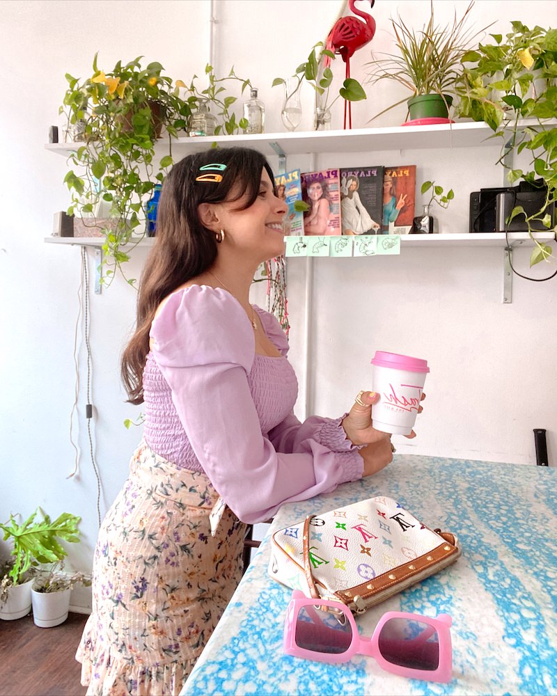 Side profile of a brunette woman who is smiling and leaning against a blue marbled coffee bar. She is wearing a lavender top and pastel multicolored floral skirt and holding a white coffee cup with a pink lid.