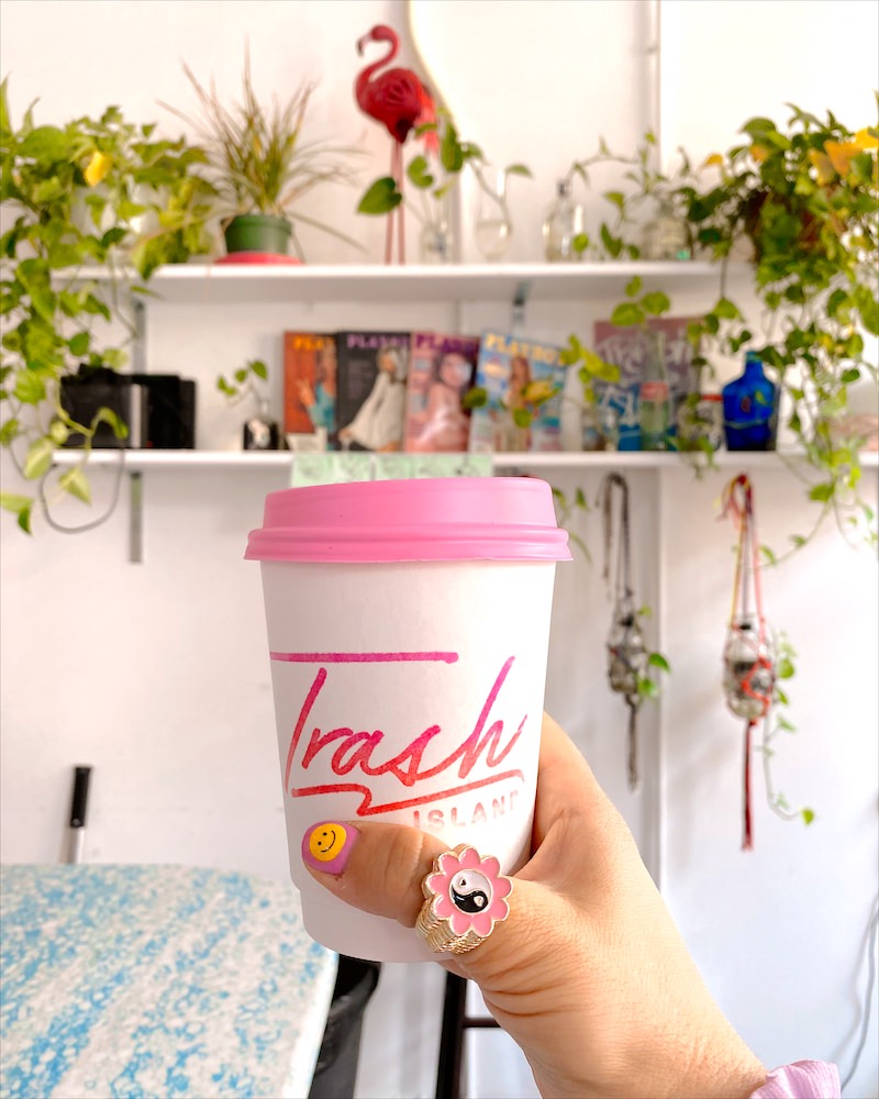 A woman's hand holding a white coffee cup that has a hot pink lid and the name of a Brooklyn Coffee Shop called "Trash Island' written in hot pink on the side. 