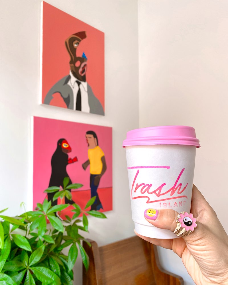 A woman's hand holding up a white and pink coffee cup in front of colorful paintings of people.