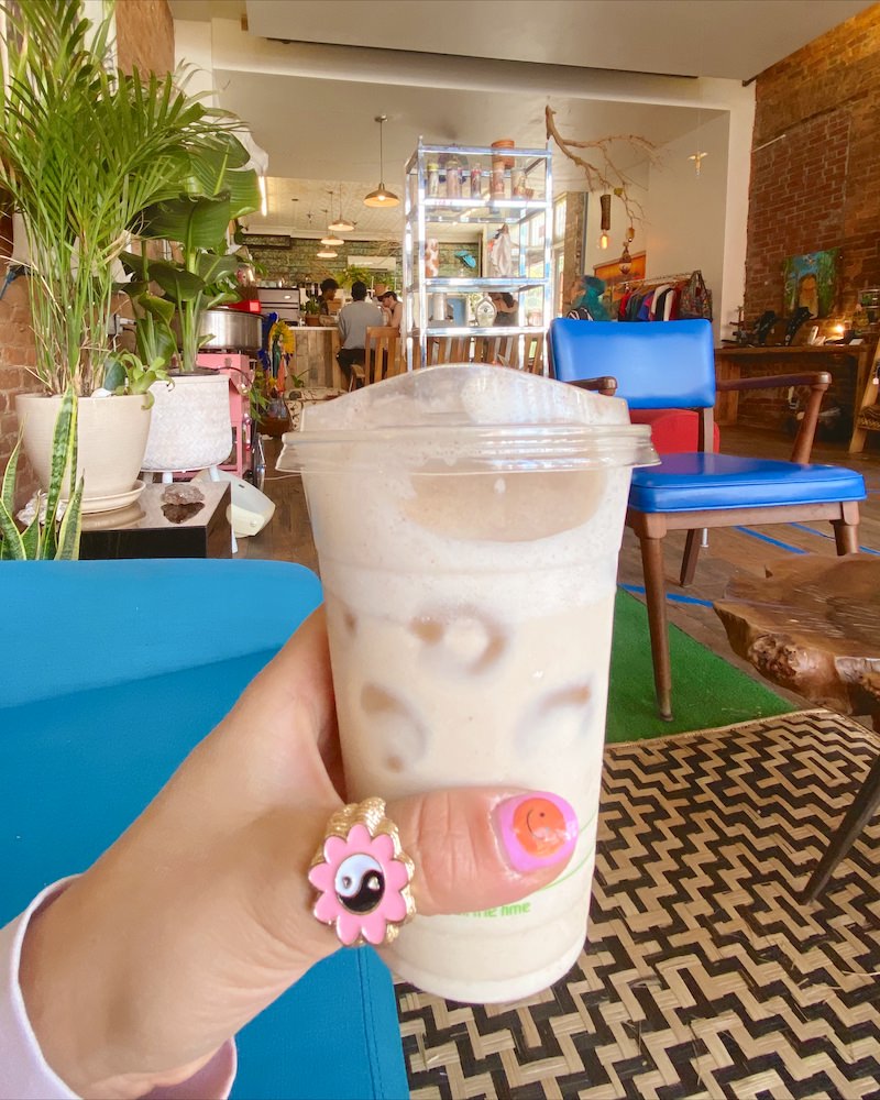 Close up photo of a woman's hand holding an iced coffee inside a Brooklyn coffee shop. She has a pink and gold yingyang statement ring on her thumb.