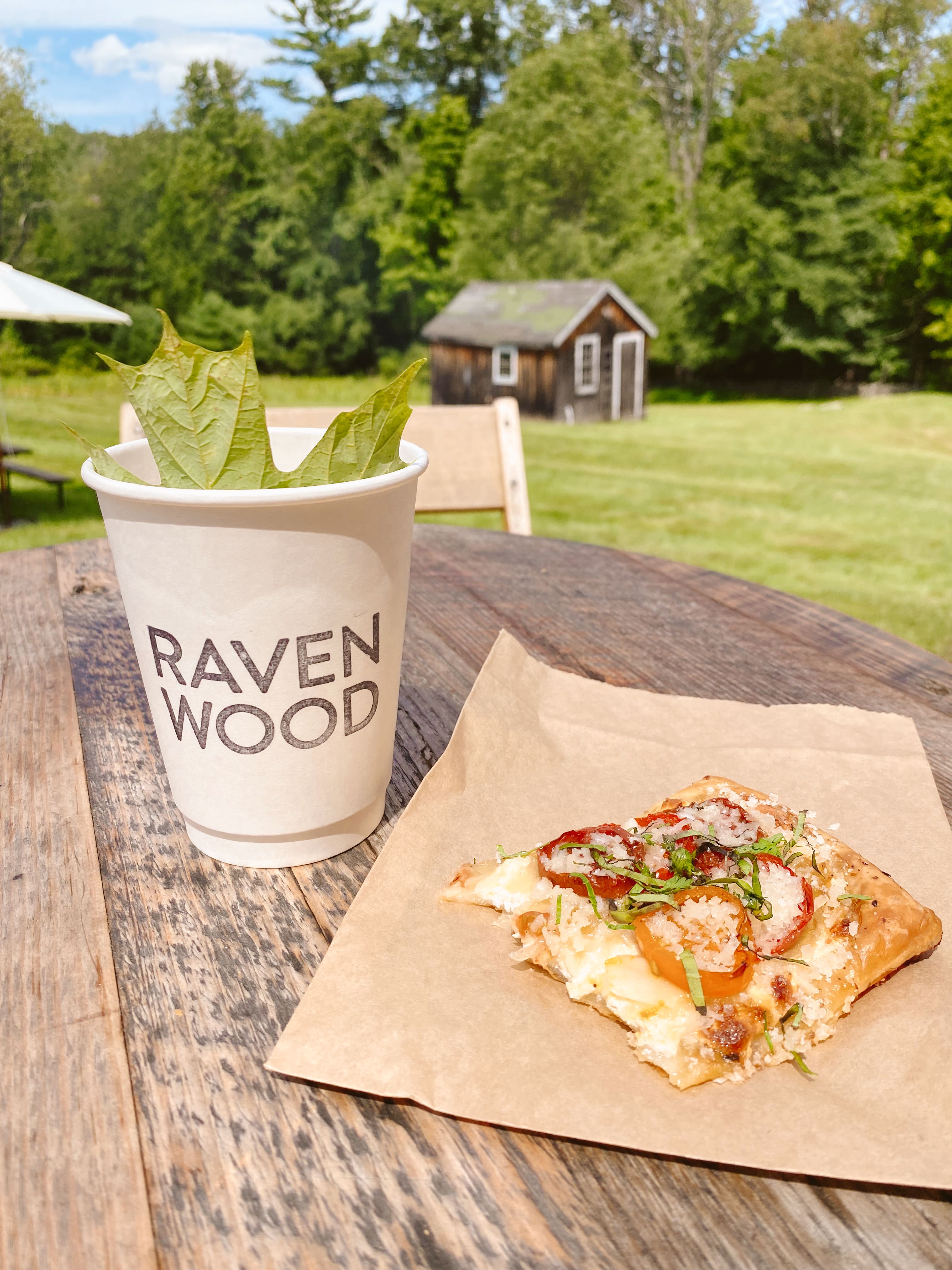 A piece of pizza and a paper coffee mug on a table with a view of a green lawn and trees at Ravenwood Barn