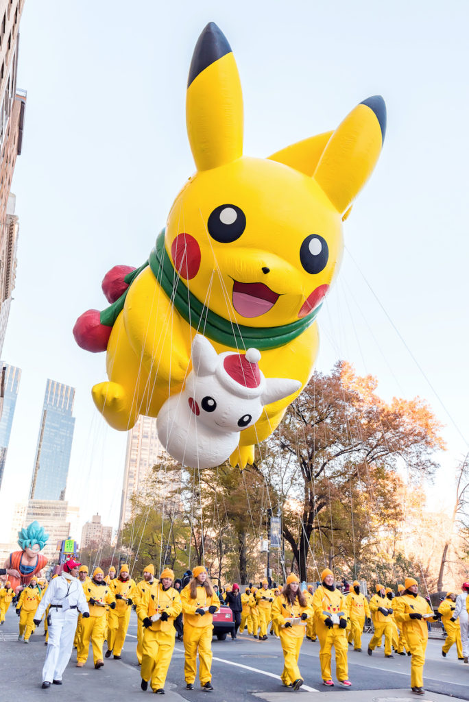 A view of the Macy's Thanksgiving Parade during fall in NYC: a large yellow Pokemon balloon is being flown above the crowd, above parade marchers wearing yellow uniforms