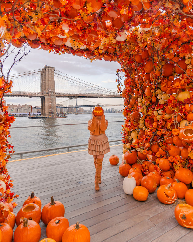 A woman wearing a beige oufit holding a jack o' lantern in front of her face. She is standing underneath an orange arch made up of hundreds of pumpkins with the Brooklyn bridge in the background seen through the pumpkin arch at Pier 17