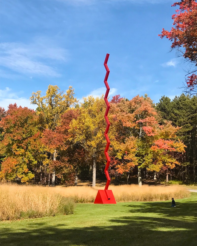 A large red modern sculpture in the Storm King Art Center garden. Behind the sculpture are trees that are turning orange and red fall colors.