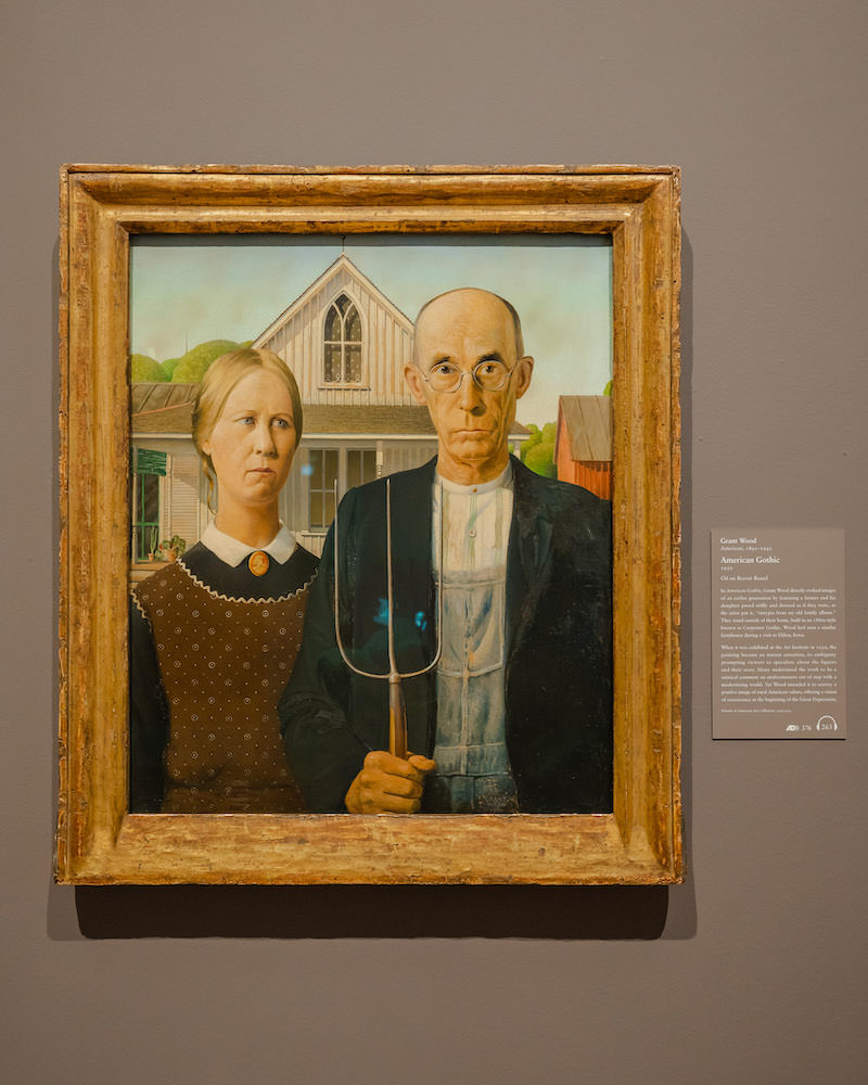 A painting called American Gothic in a museum of a serious looking woman and an older man holding in a pitchfork.