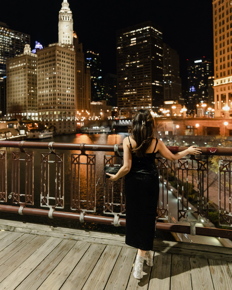 A brunette woman in a black dress is facing away from the camera with the Chicago skyline at night in the background.
