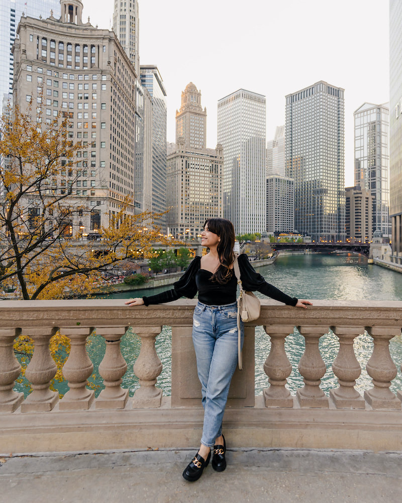 Smiling brunette woman standing in front of the Chicago River with a row of skyscrapers in the background.