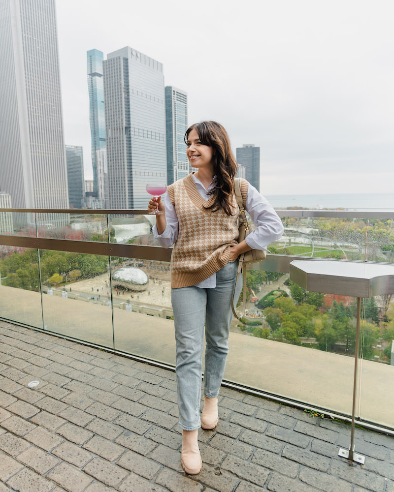 As smiling brunette woman wearing blue jeans and a tan and white checked sweater vest. She is holding a pink cocktail while standing at a rooftop cocktail bar .