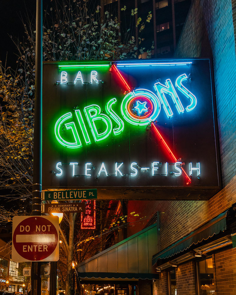 A green, bue and white neon sign that says "Bar Gibsons Steak-Fish".