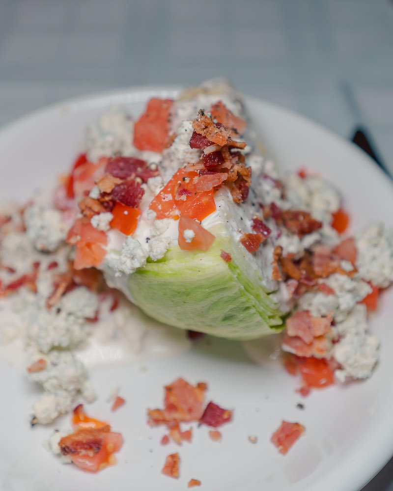 A wedge salad with romaine lettuce, tomatoes, bacon and blue cheese on a white plate. 