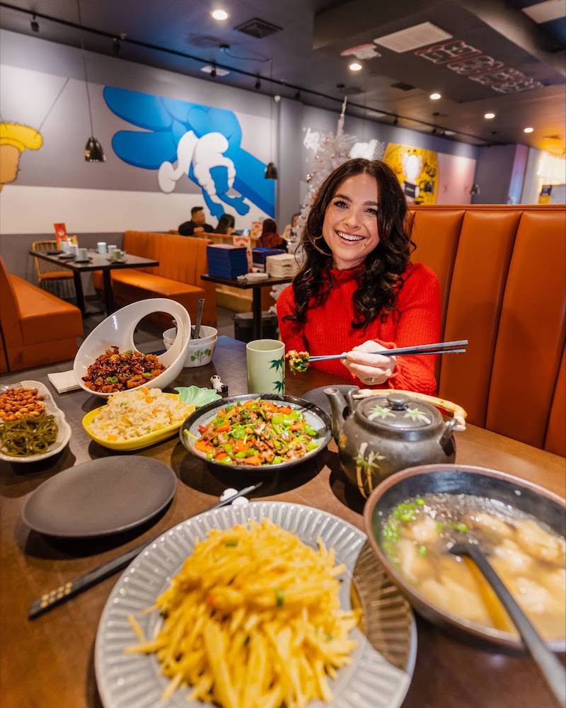 Photo of a smiling woman in a red sweater sitting in a restaurant with a large spread of Chinese food on the table.