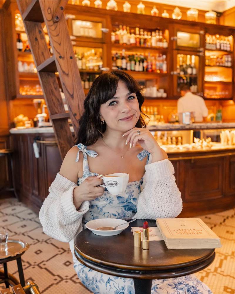 A brunette woman sitting in a cafe in a blue and white floral print dress. She is smiling and holding a cappuccino.