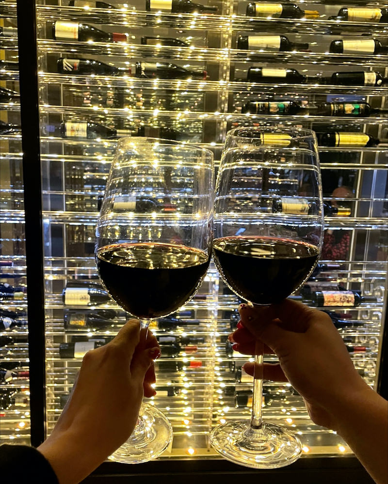 Two women's hands holding glasses of red wine in front of a display of wine bottles.