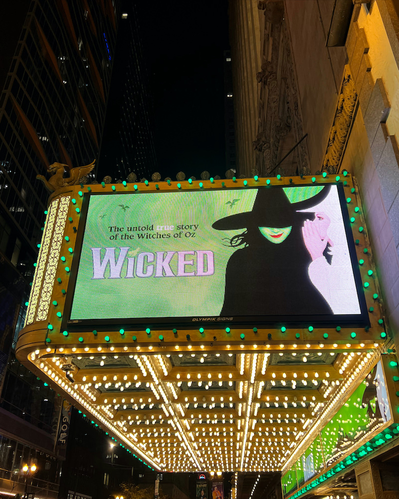 A lit up gold marquee with a white witch whispering into a green witches's ear. The sign reads "the untold true story of the Witches of OZ - Wicked"