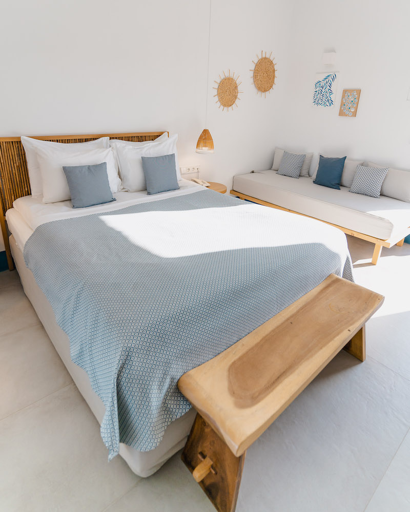The interior of a hotel room at Milos Breeze Boutique Hotel that has a wooden bed with blue and white bed linens and a small couch with blue and white cusions.