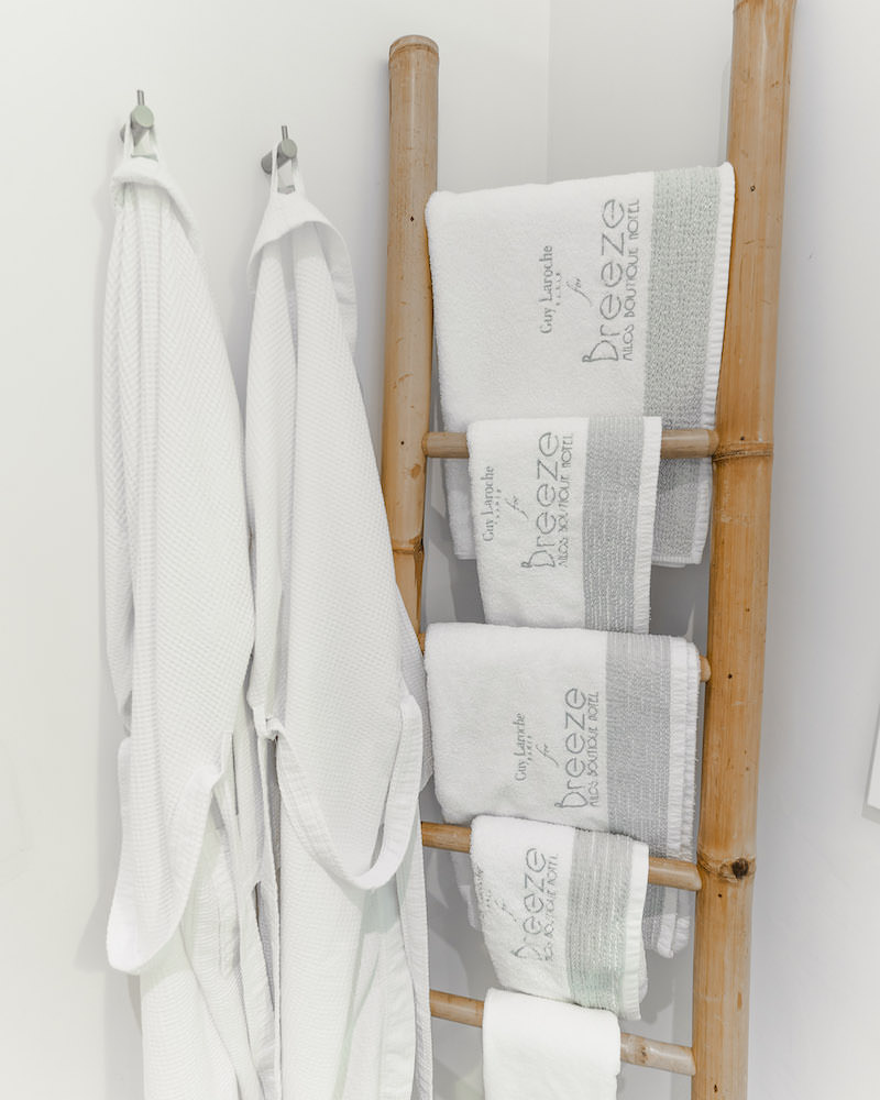 A wooden towel rack with white towels on it and 2 white hotel bathrobes hanging from the walls on hooks.