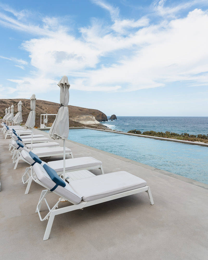 A row of sun loungers with blue and white cushions that face a big hotel infinity pool and the ocean.