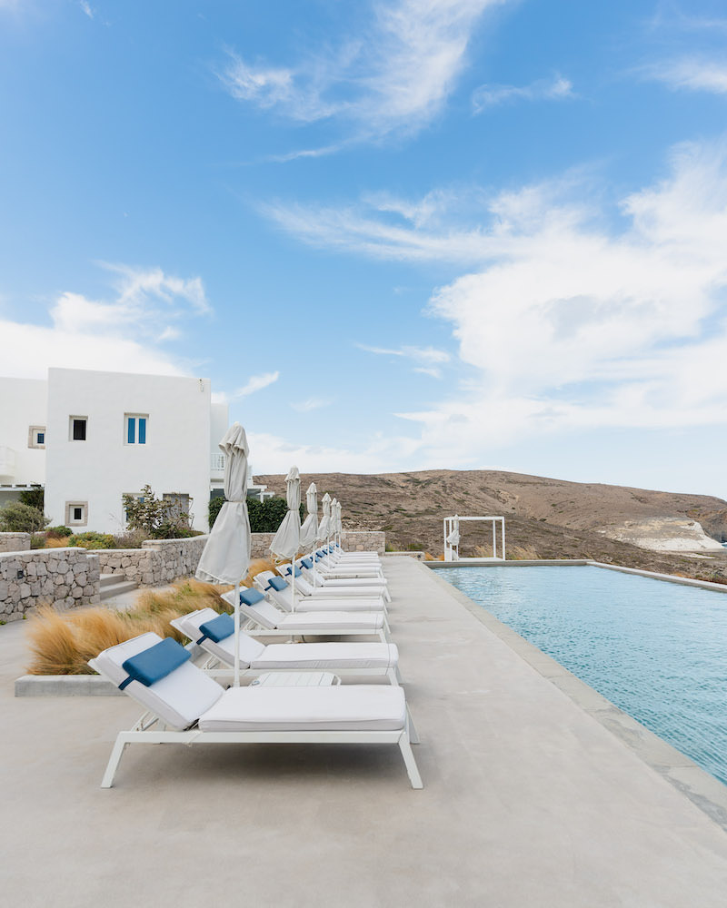 A row of white lounge chairs with white and blue pillows that face a large infinity pool.